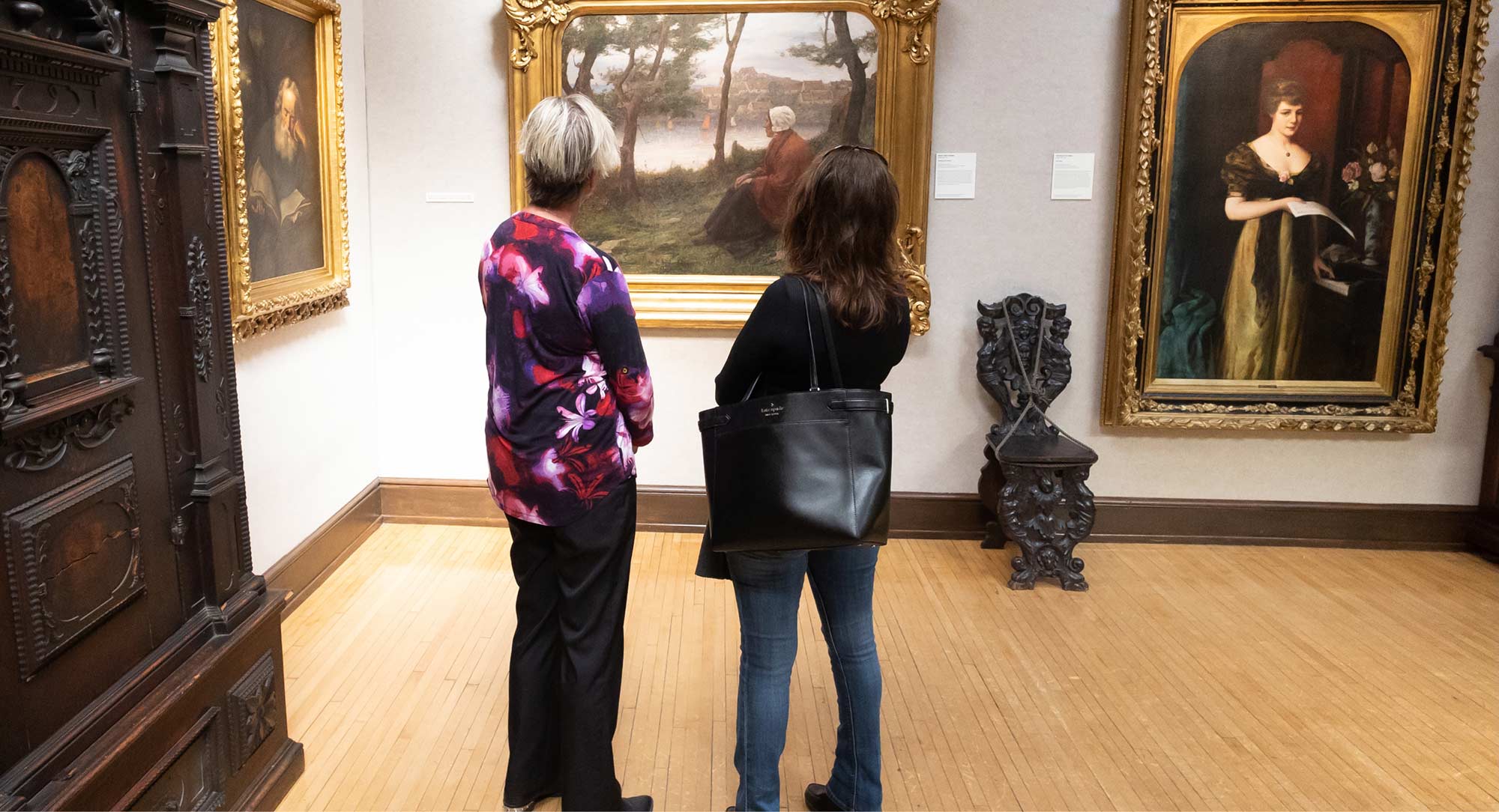 Two women in the galler looking at a painting of a woman sitting under a tree wearing a white bonnet and a red cape.