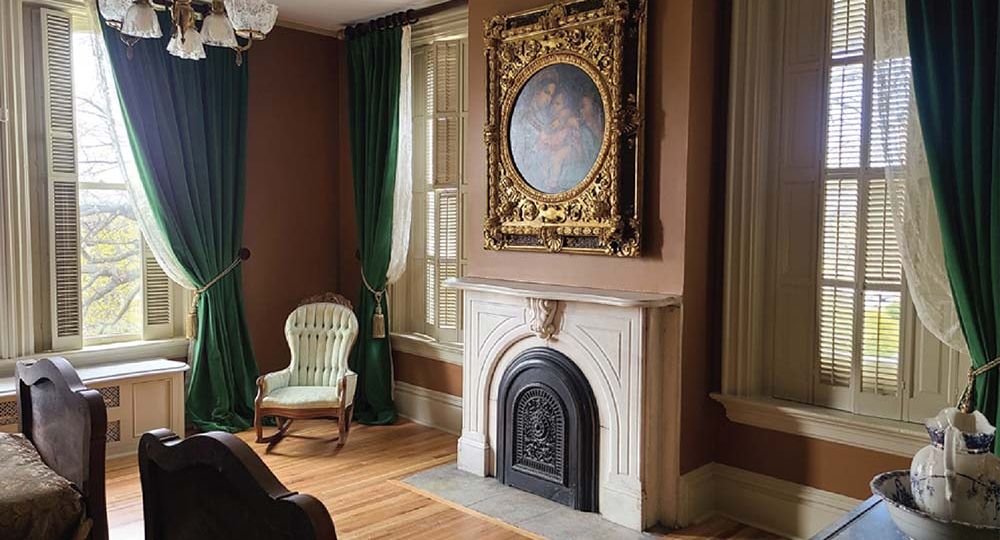 An interior view of a bedroom in the mansion, with an ornate framed painting over a fireplace. Heavy curtains are pulled back with ties revealing wooden shutters.