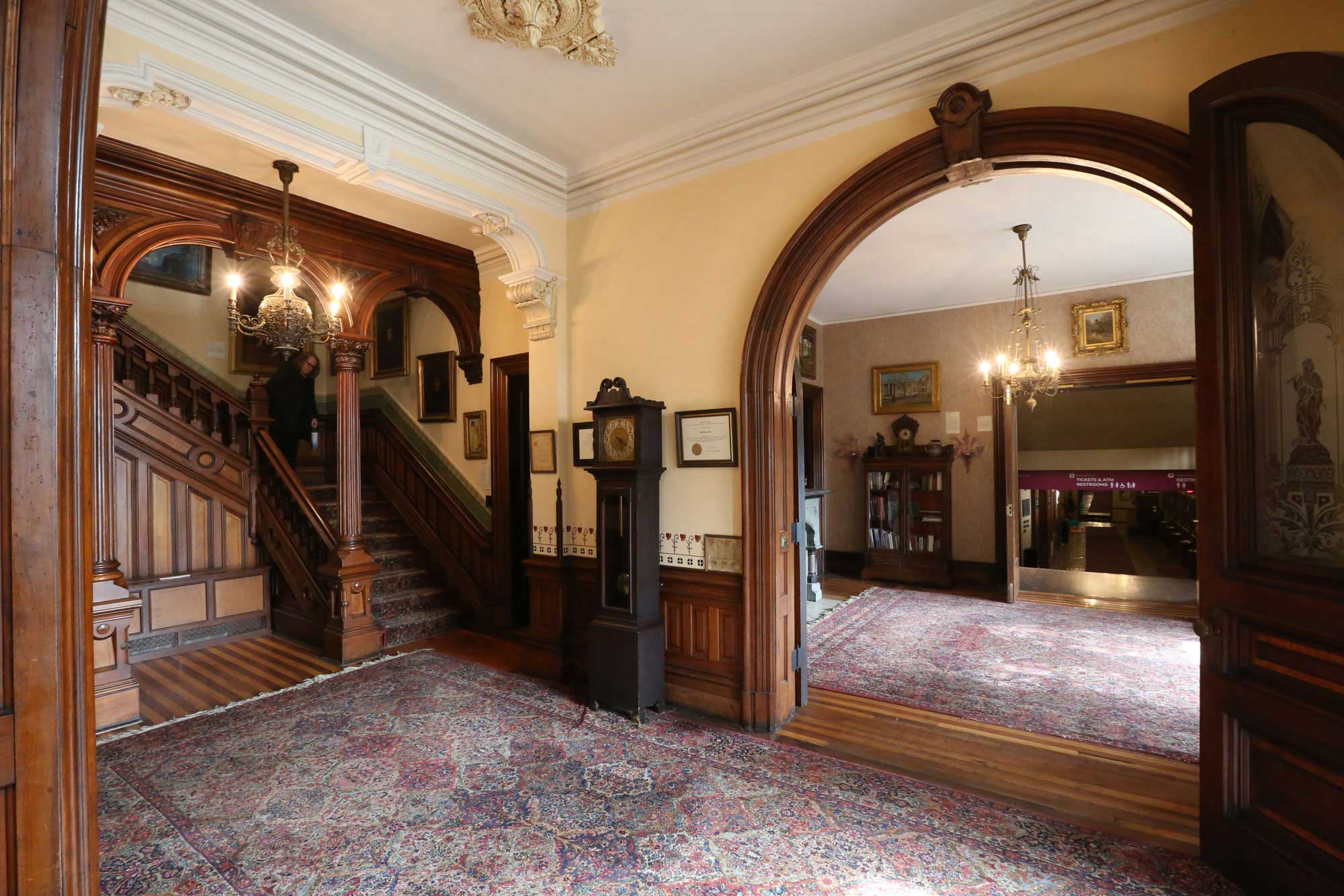 An interior view of the mansion with a large arched double doorway, a grandfather clock, and an ornately carved set of wood columns that frame a staircase.