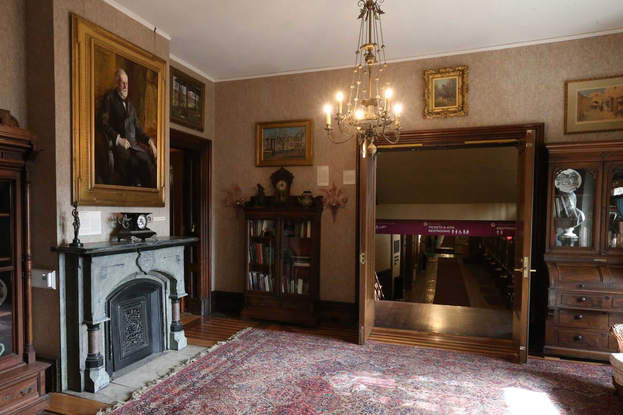 An interior view of the mansion with a dark stone fireplace with a portrait of Hoyt Sherman above the mantle. Double doors are open to reveal the hallway that leads to the theater.