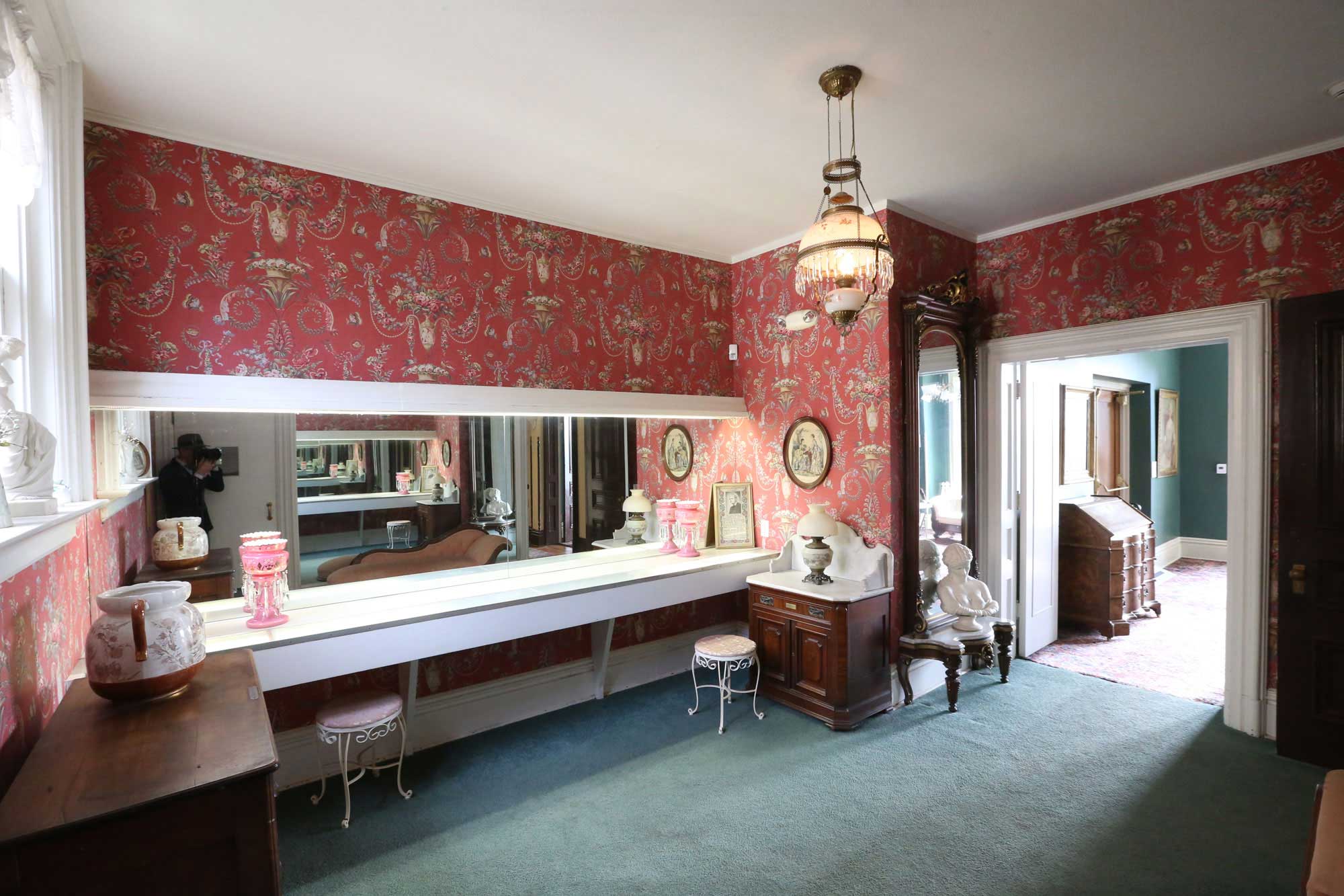An interior of the ladies powder room with red floral wallpaper, a long mirrored shelf with seating, and a chandelier.